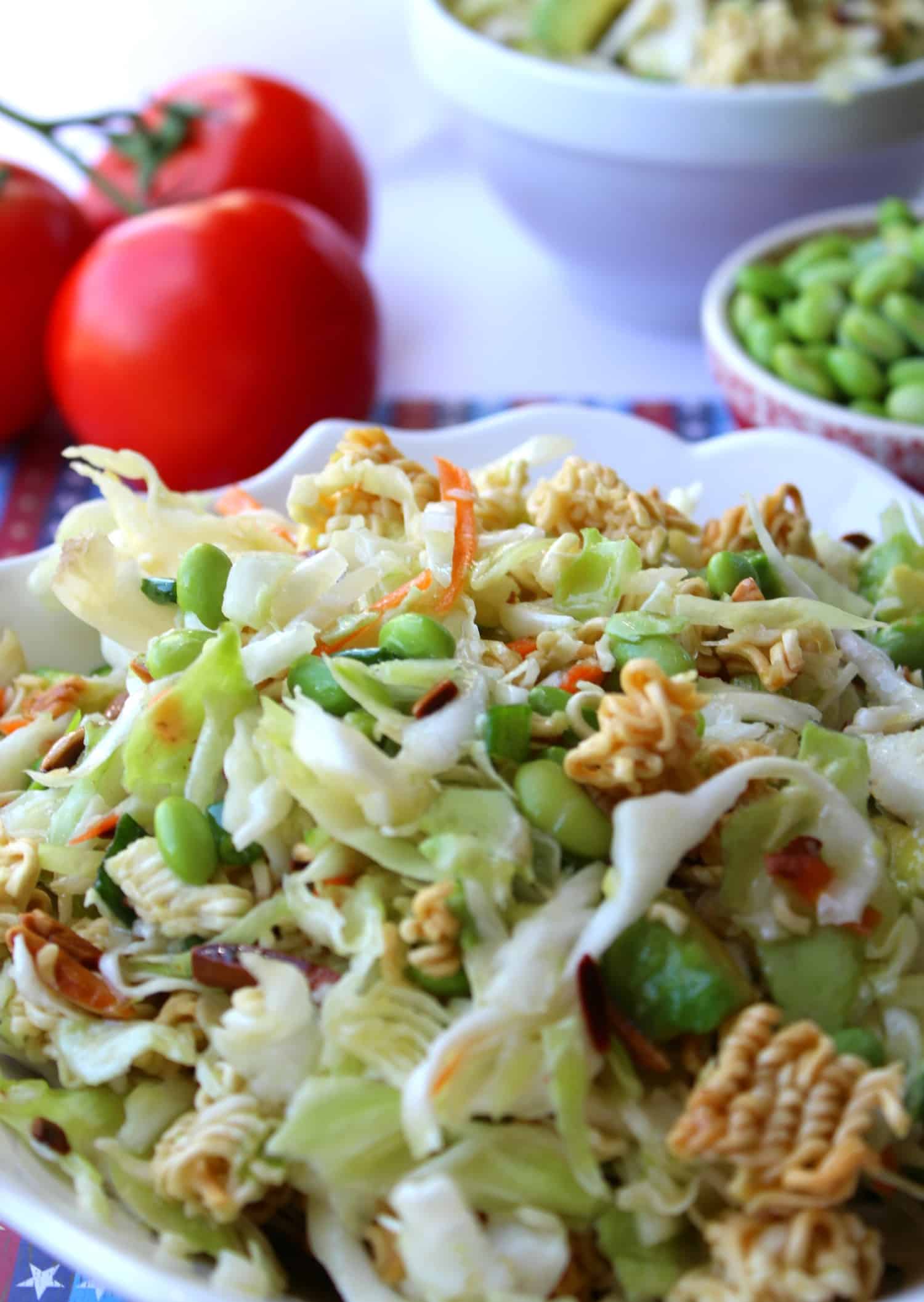 A bowl of cabbage salad with edamame and crunchy ramen noodles, with tomatoes and more salad in the background.