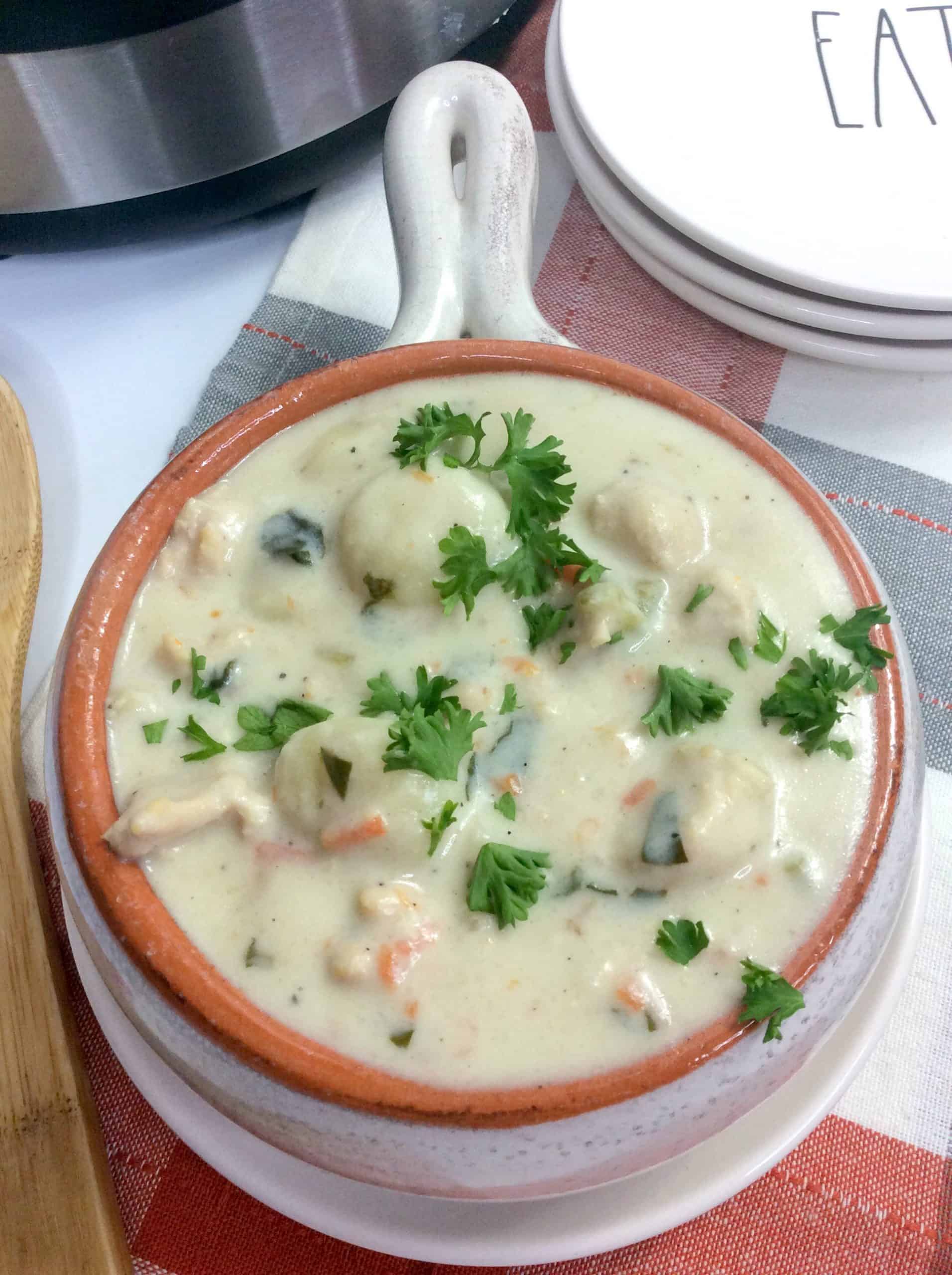 A bowl of creamy Instant Pot Chicken Gnocchi Soup with vegetables, chicken pieces, and parsley garnish sits on a red and white cloth next to a stack of white plates.