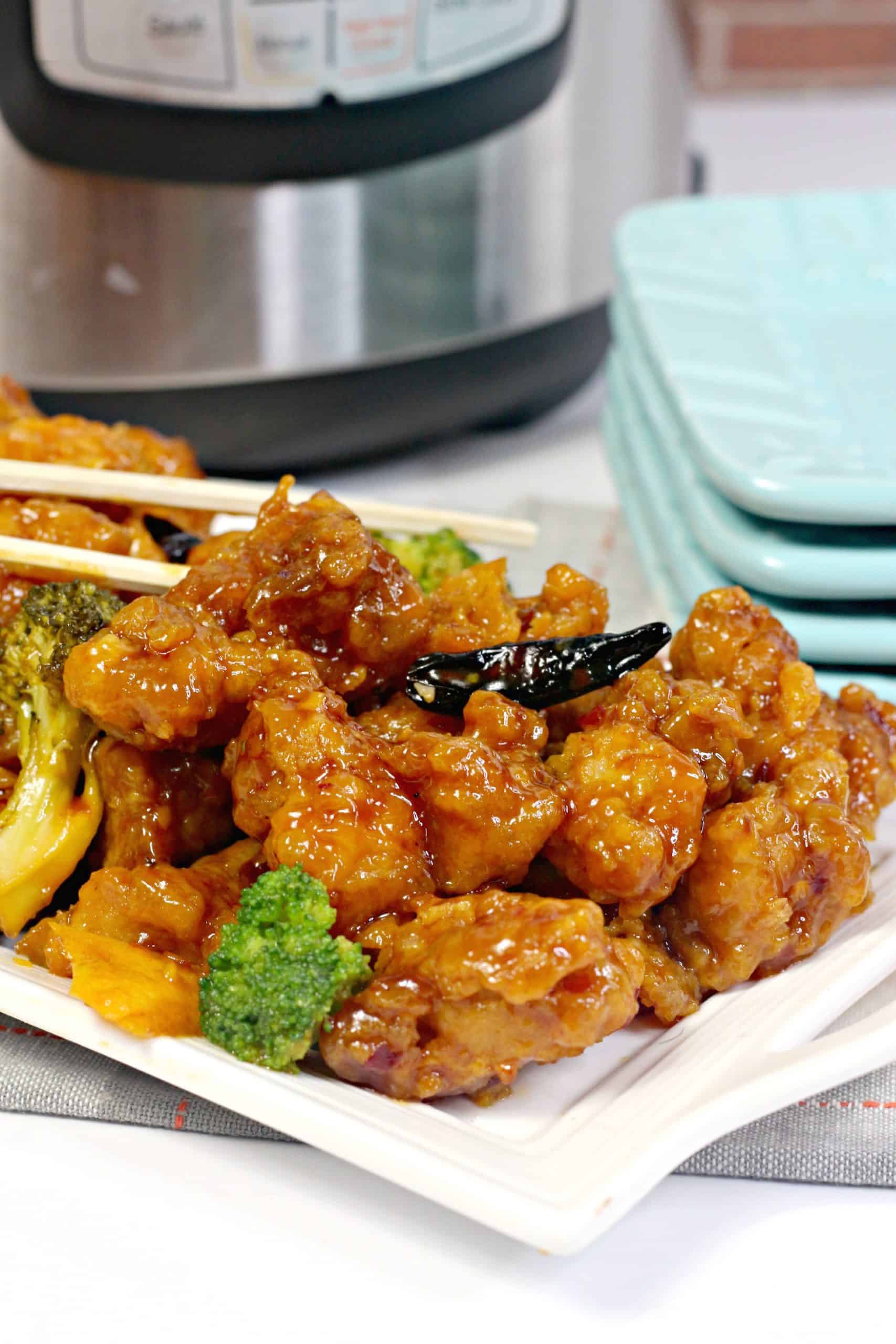 Plate of orange chicken with broccoli, garnished with dried chili peppers, in front of a stack of blue plates and an electric pressure cooker.