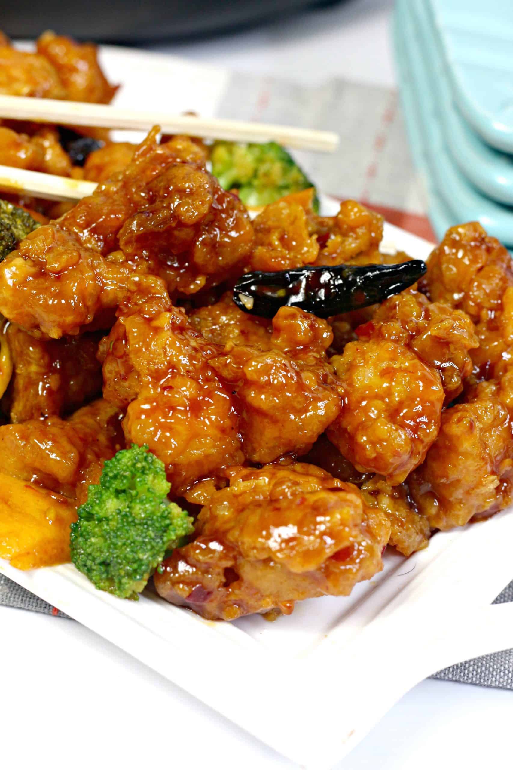 Close-up of crispy, saucy orange chicken pieces with broccoli, served on a white plate with chopsticks.