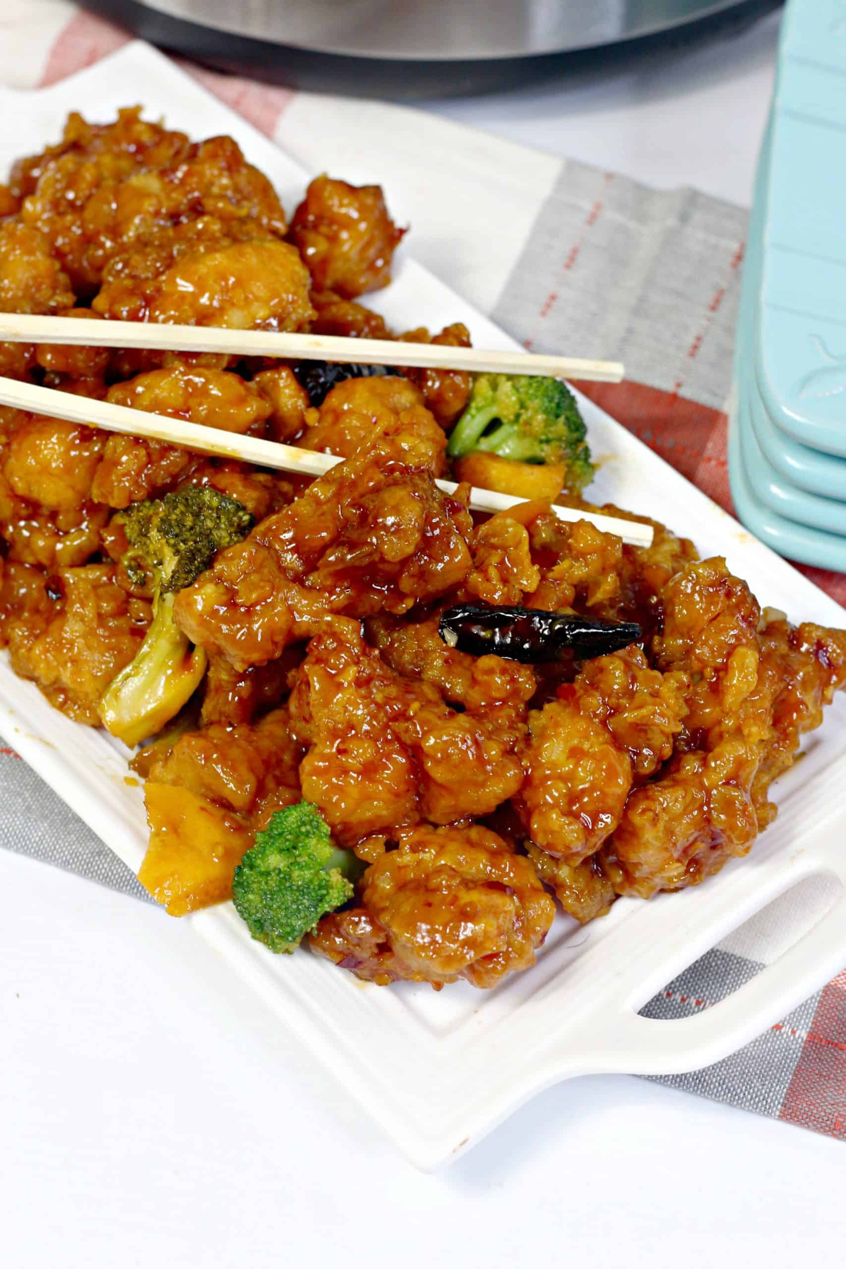 A rectangular white plate with pieces of glazed orange chicken, broccoli florets, and chopsticks resting on top.
