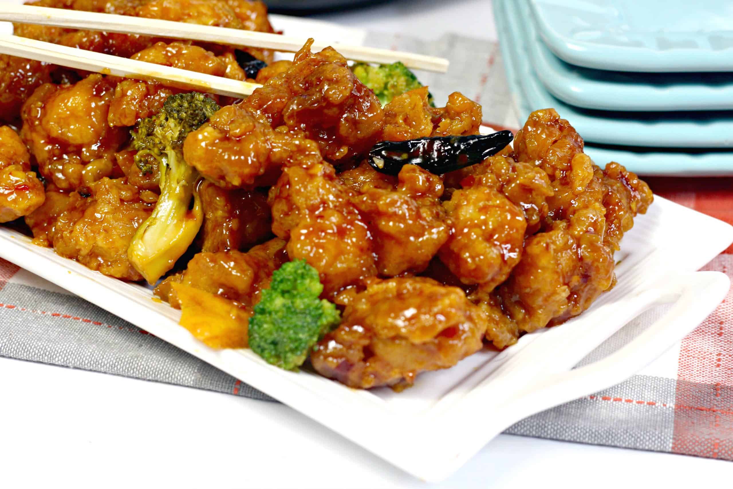 A rectangular white plate with crispy glazed chicken pieces, broccoli florets, and dried chili peppers, with chopsticks resting on top.