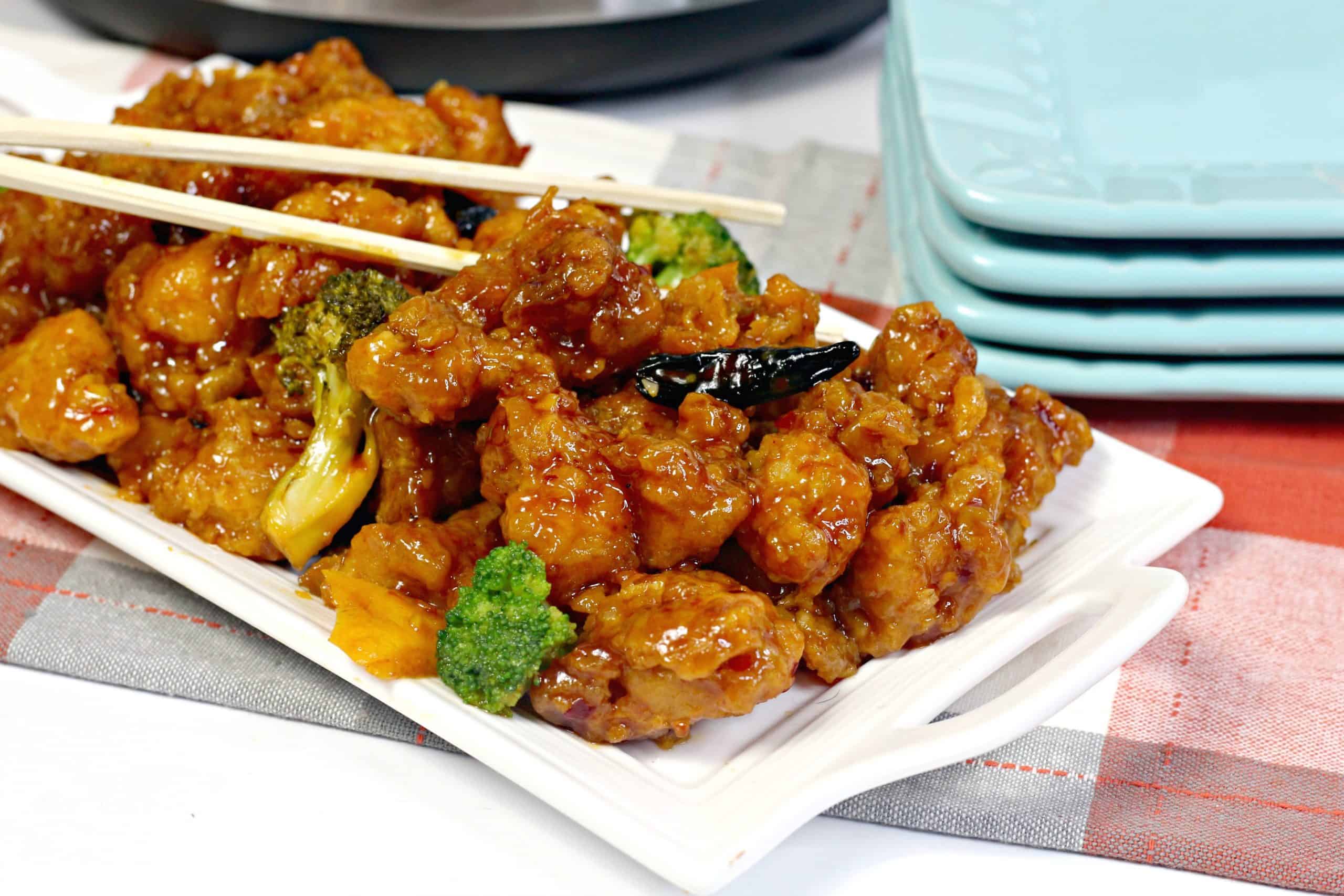 A rectangular white plate with General Tso’s chicken, broccoli florets, dried chili peppers, and chopsticks on top; stacked blue plates are in the background.