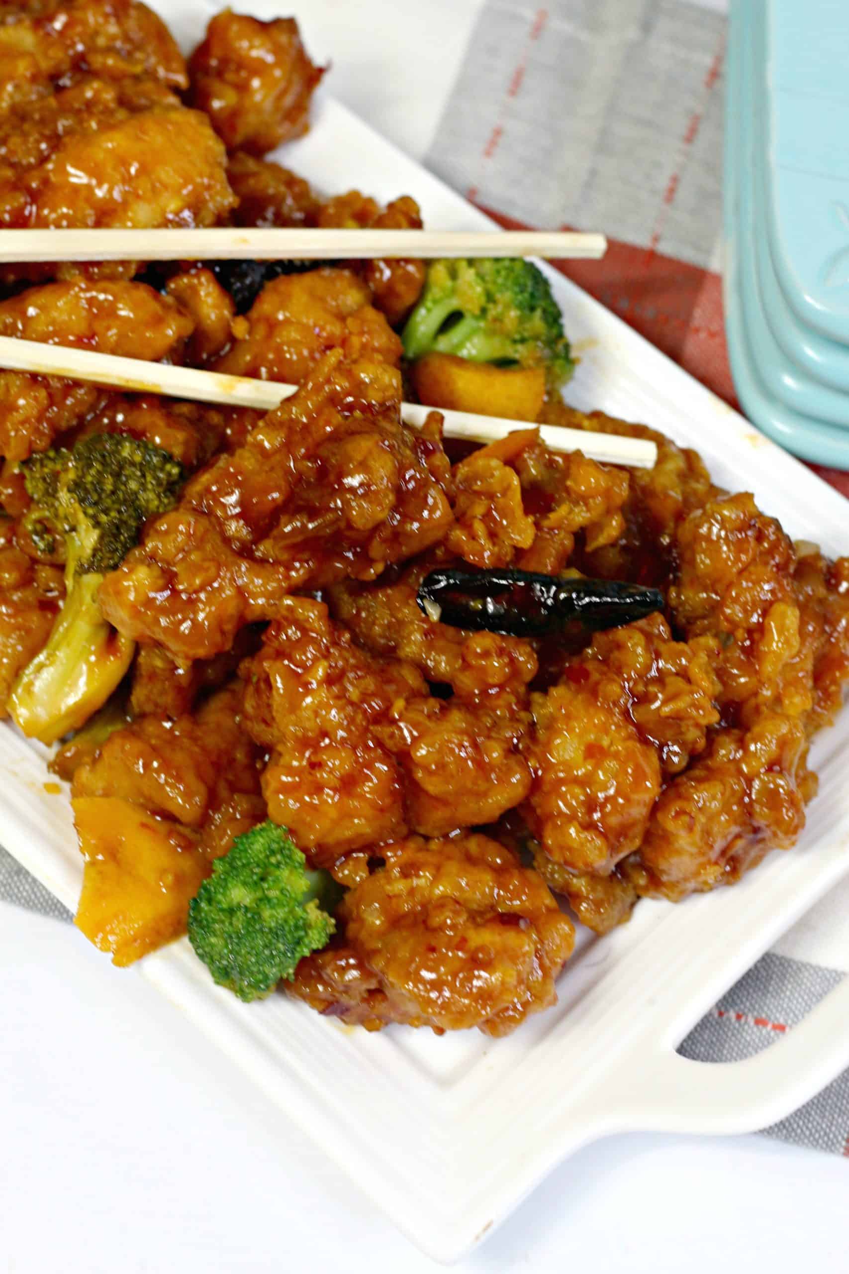 A rectangular white plate with crispy, sauced chicken pieces, broccoli florets, and chopsticks resting on top.