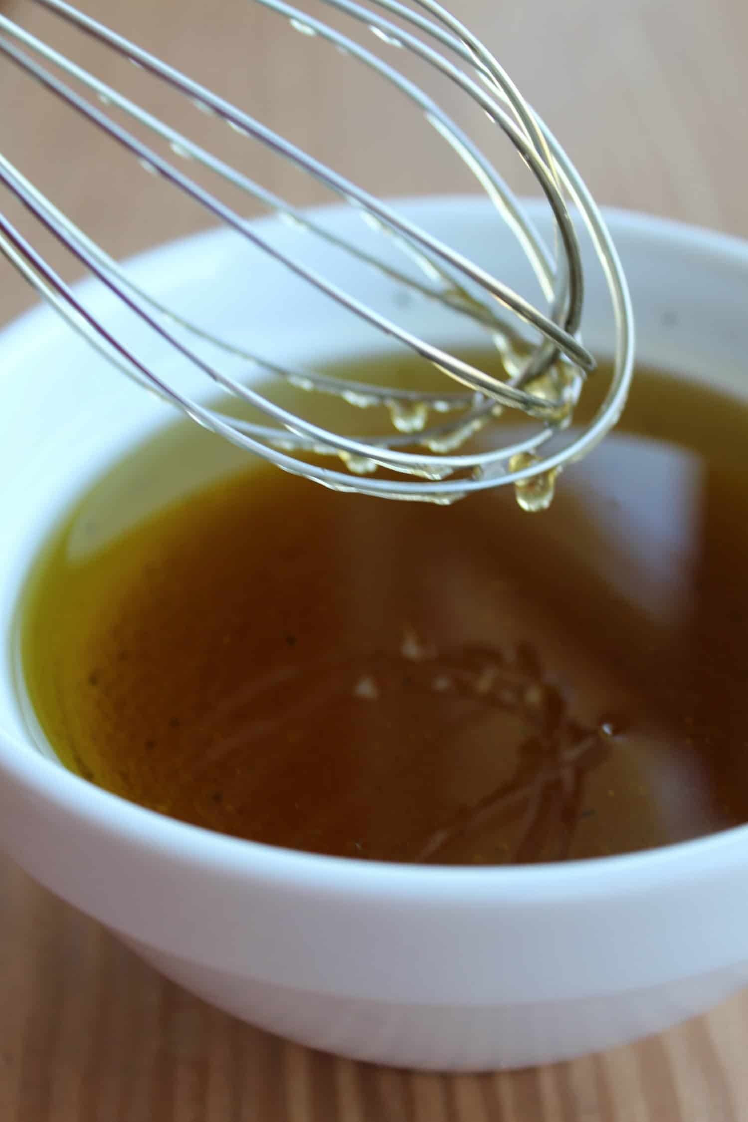 A metal whisk with liquid drips over a white bowl filled with a golden-brown sauce or dressing on a wooden surface.