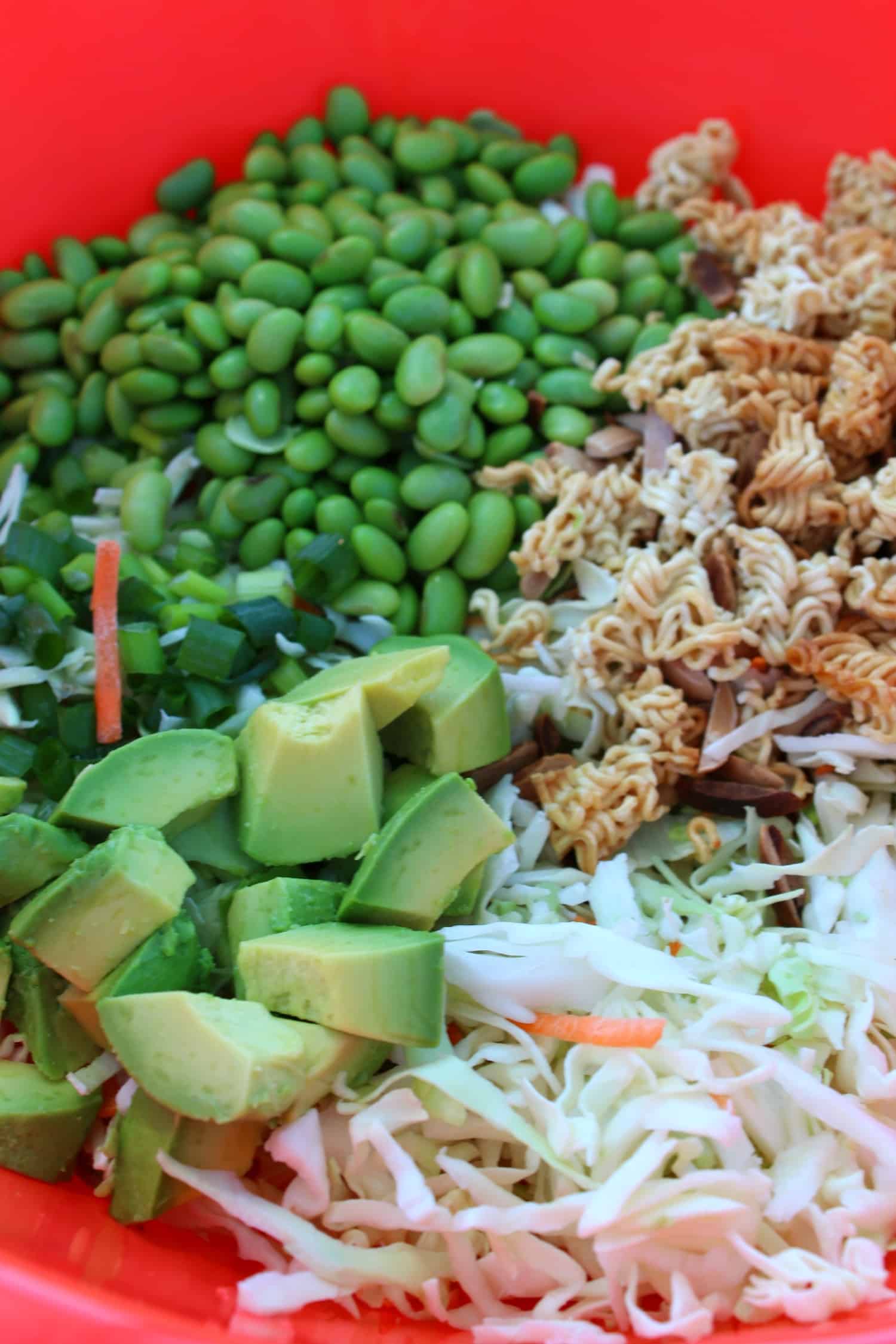 A red bowl containing chopped cabbage, sliced avocado, green onions, shelled edamame, shredded carrots, and broken pieces of dry ramen noodles.