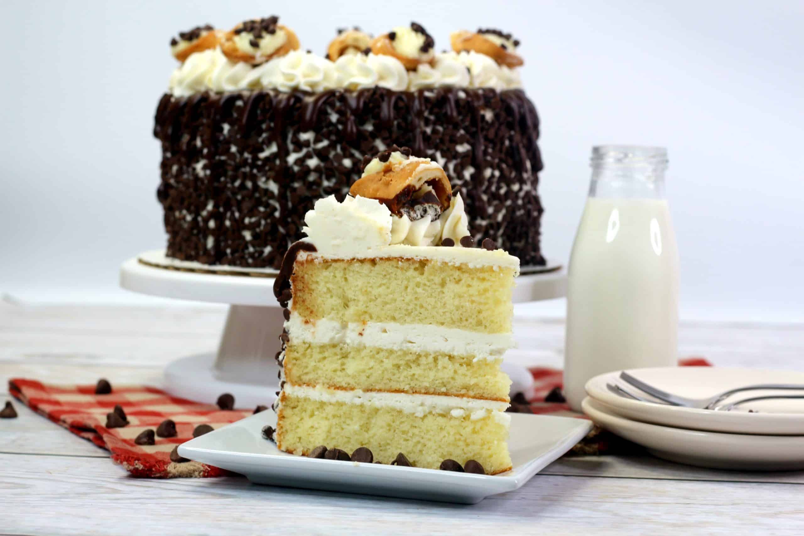 A slice of layered vanilla cake with white frosting and chocolate drizzle sits on a plate in front of a whole decorated cake, a bottle of milk, and a stack of plates.