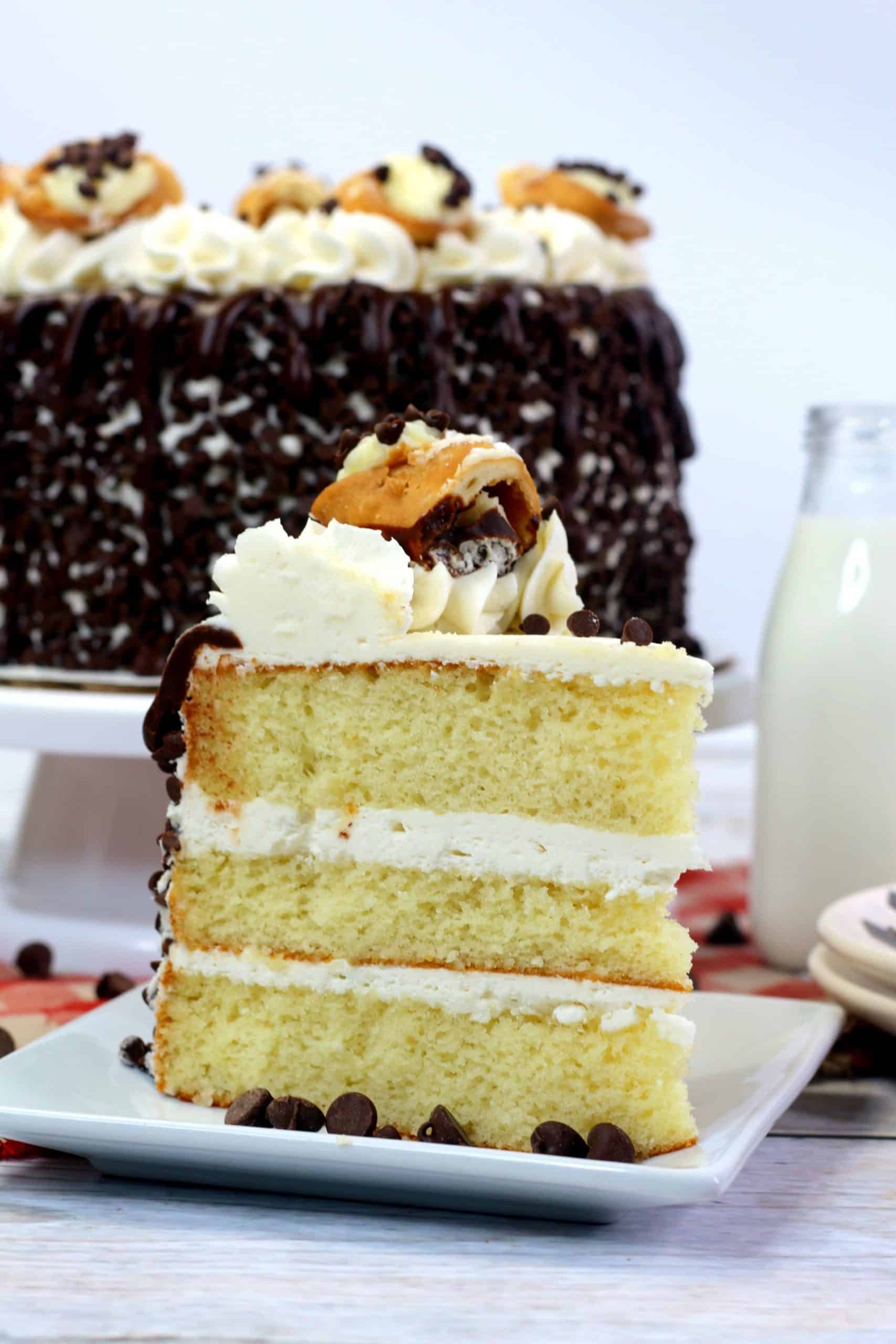 A slice of layered vanilla cake with white frosting, chocolate chips, and cream topping sits on a plate; the whole cake and a bottle of milk are in the background.