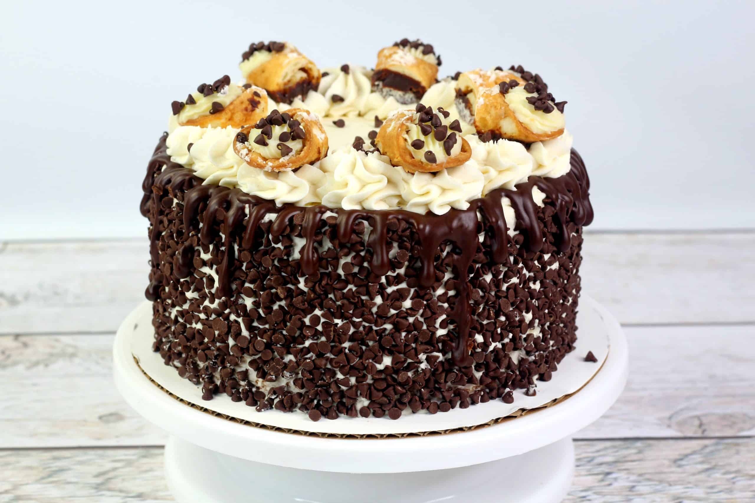 A round chocolate cake with white frosting, chocolate drizzle, mini chocolate chips on the sides, and cream puffs topped with chocolate chips on top, displayed on a white cake stand.