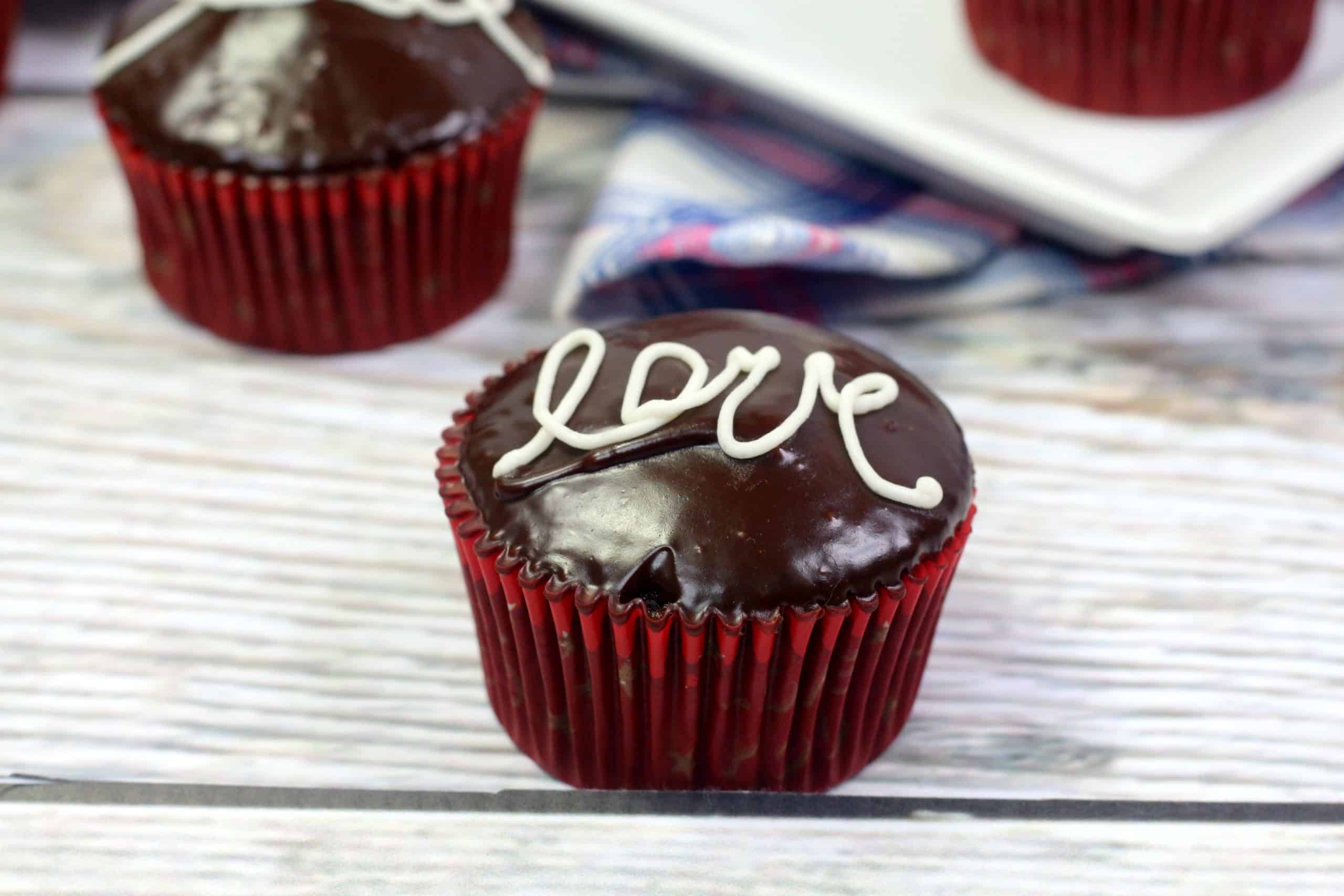 A chocolate cupcake with glossy icing, decorated with the word love written in white icing, in a red paper liner on a wooden surface.