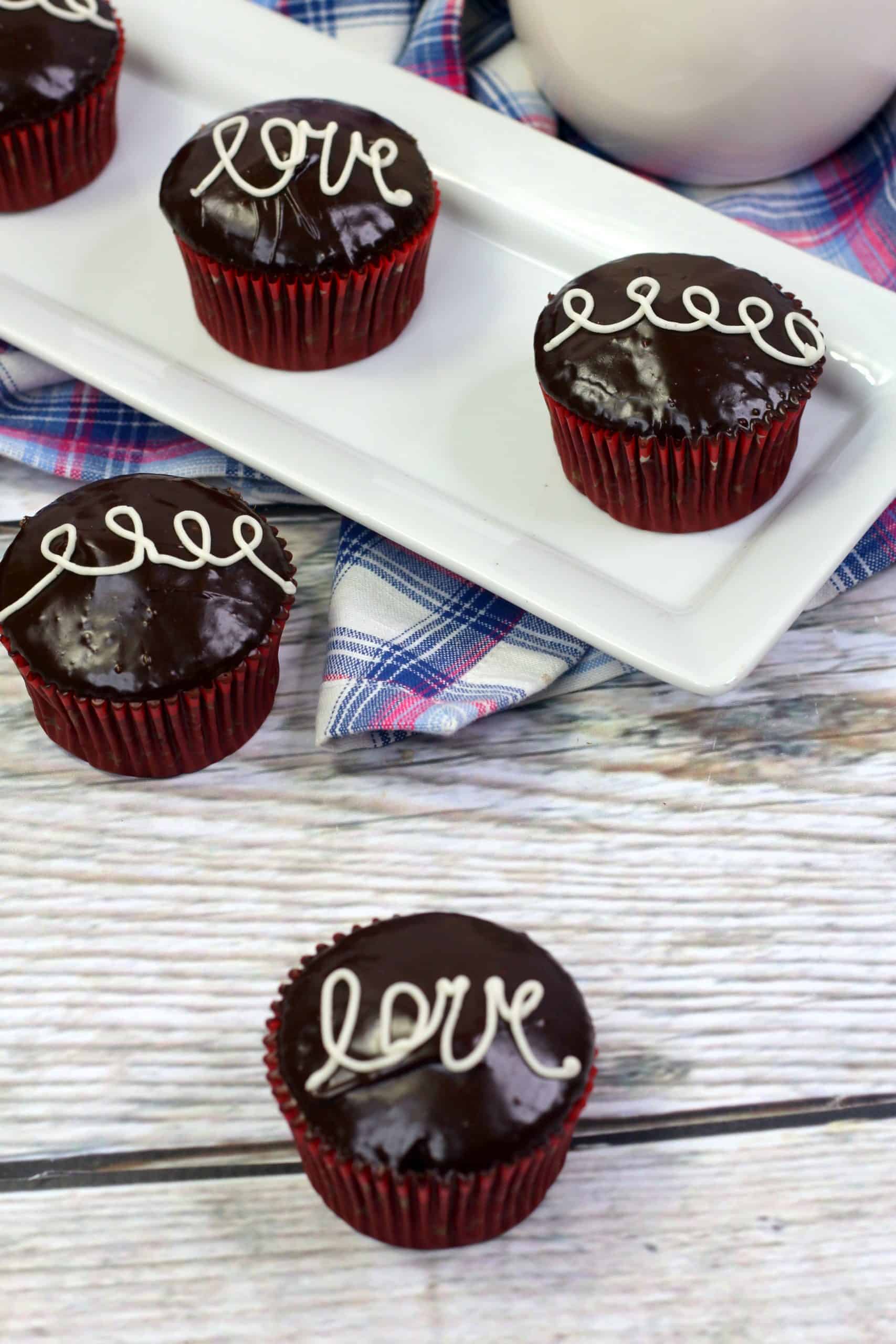 Four chocolate cupcakes with glossy icing and the word love or a swirl piped in white, arranged on a tray and table with a plaid cloth in the background.