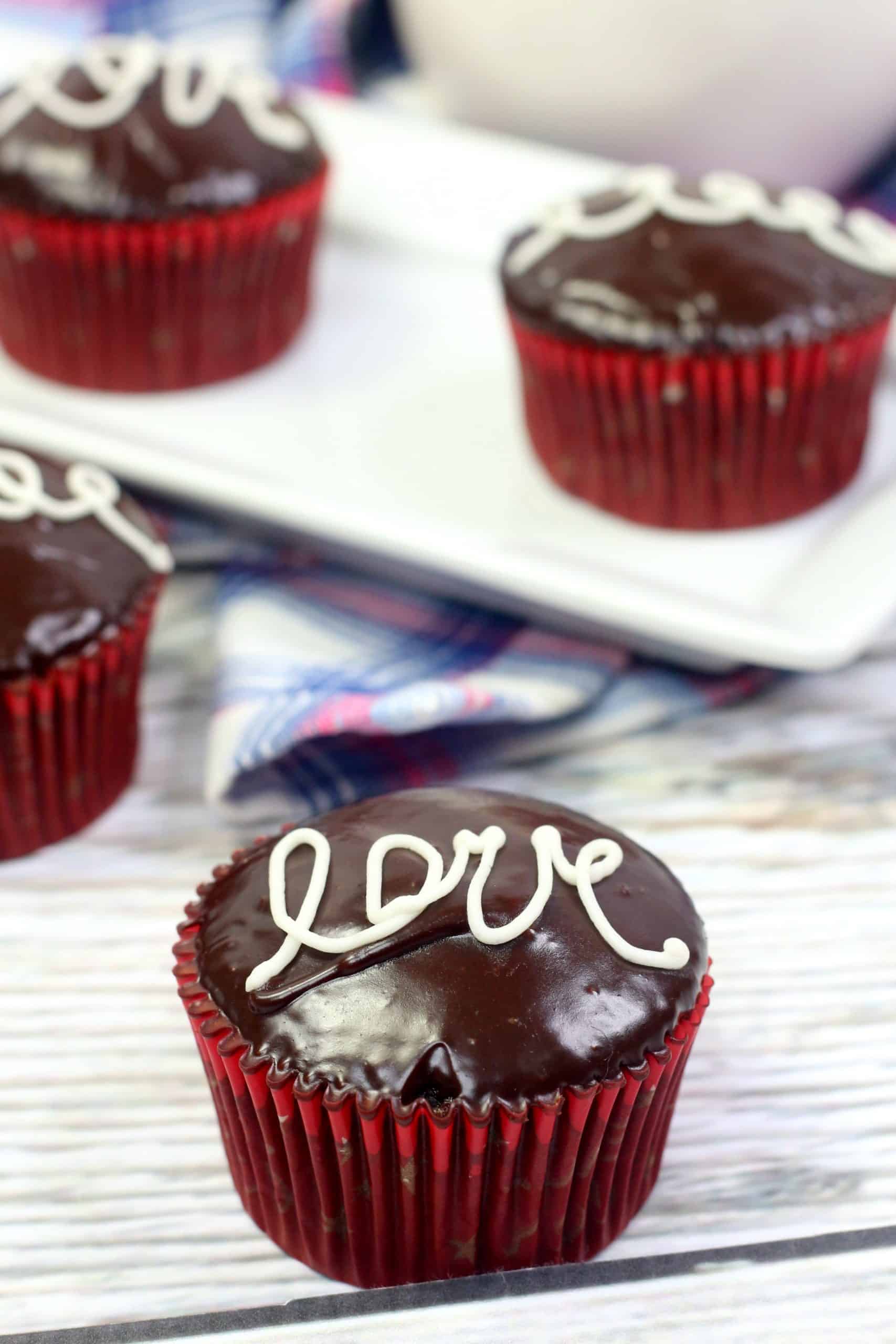 Chocolate-glazed cupcake in red wrapper with the word love written in white icing, placed on a light wooden surface near other similar cupcakes.