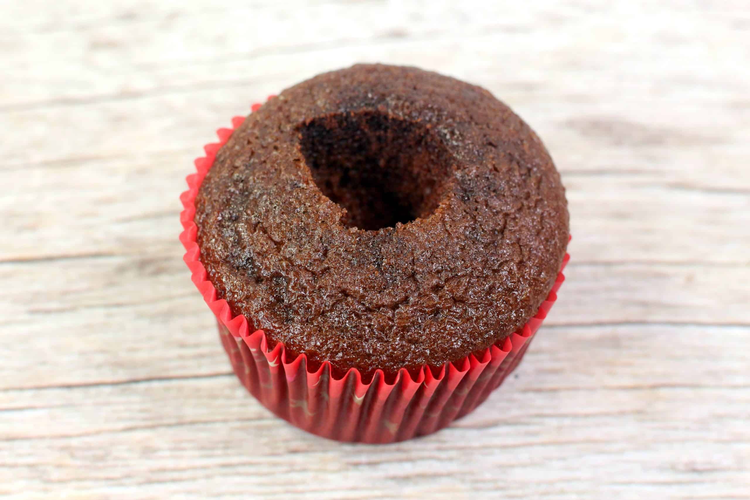 A chocolate cupcake with a hole in the center, sitting in a red paper liner on a wooden surface.