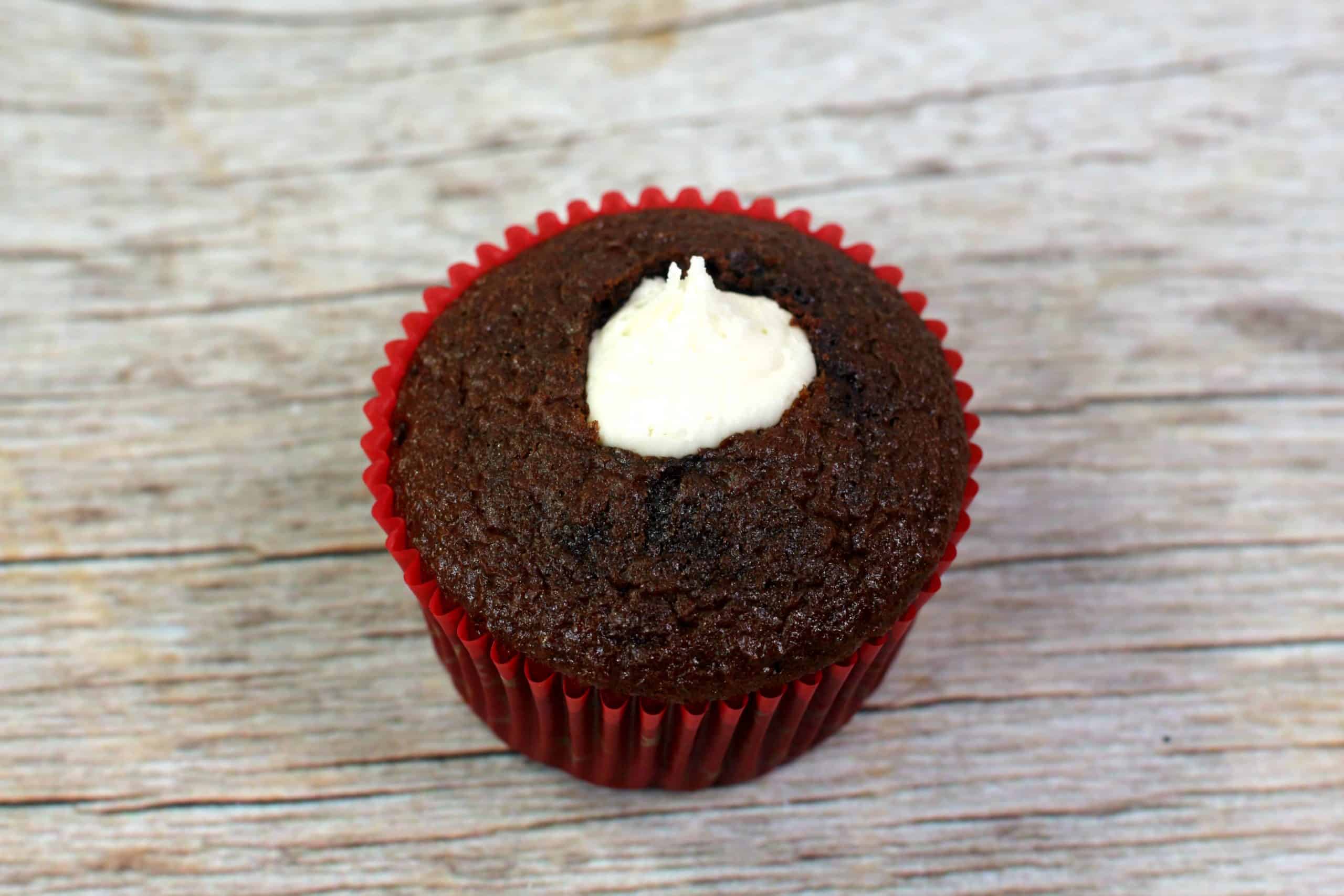 A chocolate cupcake in a red paper liner with a dollop of white cream filling on top, placed on a wooden surface.