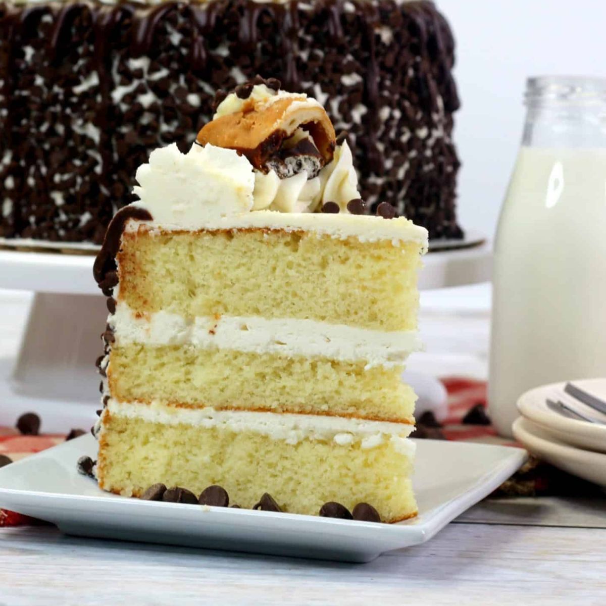 A slice of vanilla layer cake with white frosting and chocolate chips on a plate, with a whole cake, a glass of milk, and stacked plates in the background.