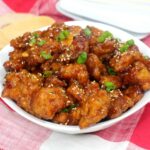 A white bowl filled with glazed, crispy General Tso Chicken pieces garnished with sesame seeds and chopped green onions, placed on a red and white cloth.