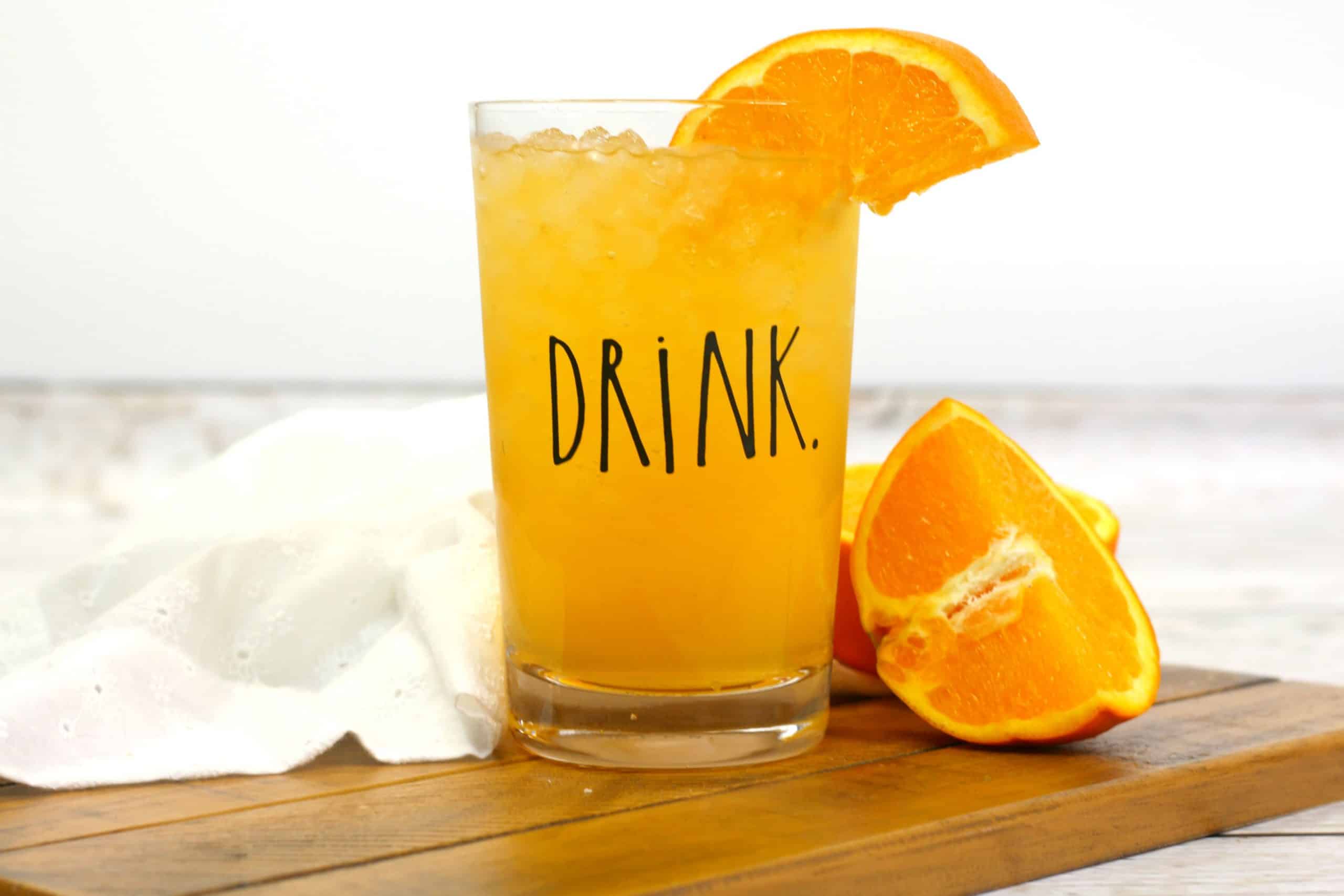 energy drink in a clear glass with a drink label on it, with a slice of orange in the drink and oranges next to it.
