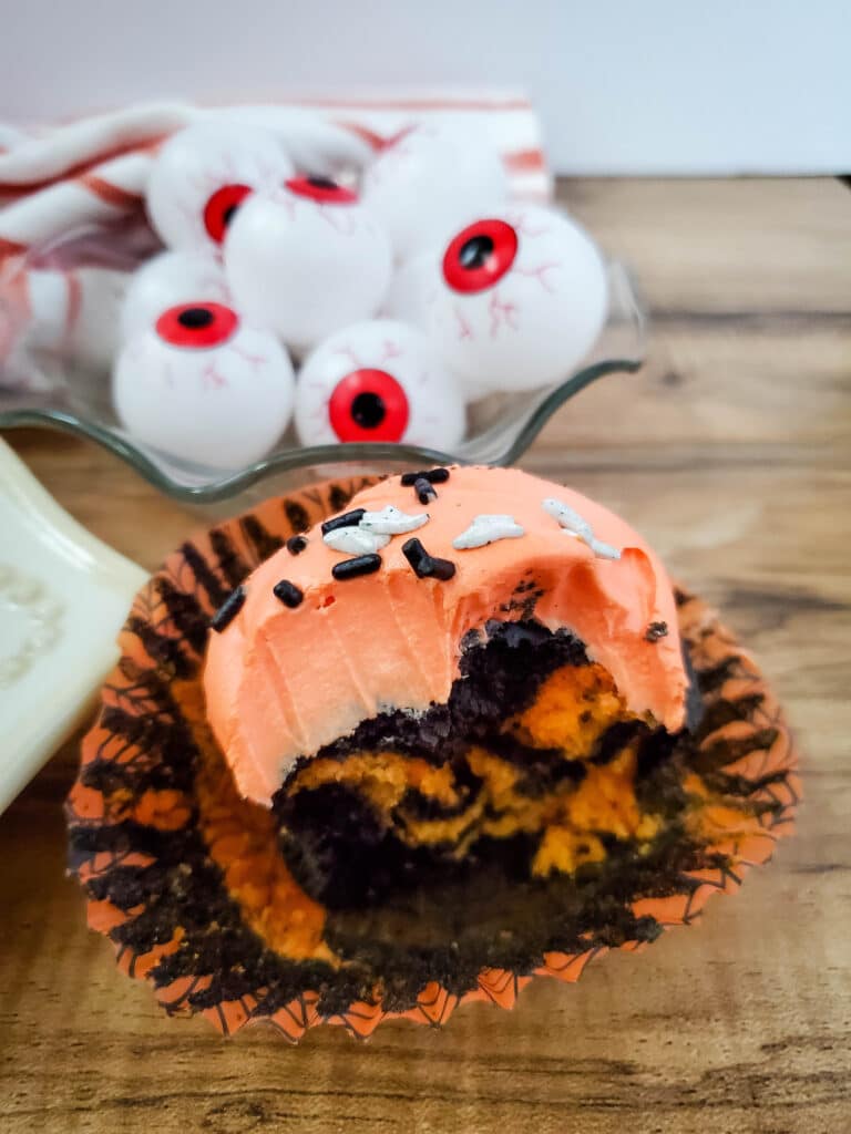 Vertical image of a tie dye cupcake with orange frosting, with a bite missing. A bowl of eye balls in the background.