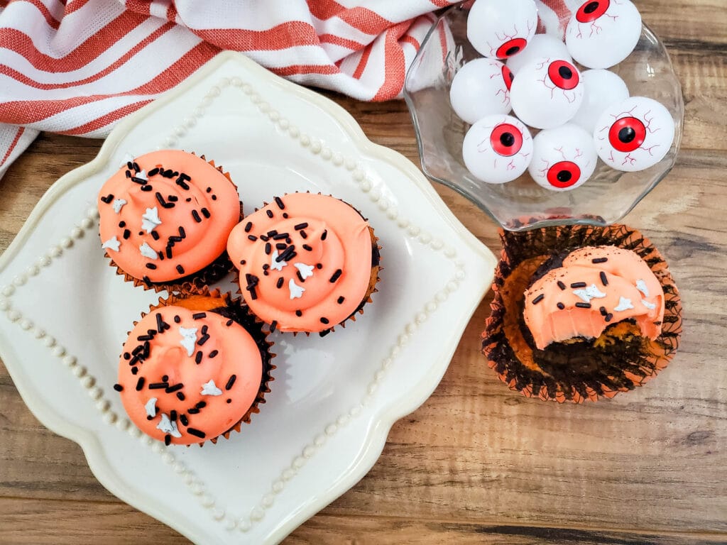 birds eye image of three orange and black tie dye cupcakes on a plate, with another cupcake with a bite missing next to the plate.