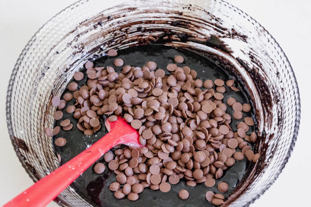 brownie batter with chocolate chips in a large glass mixing bowl with red rubber spatula.