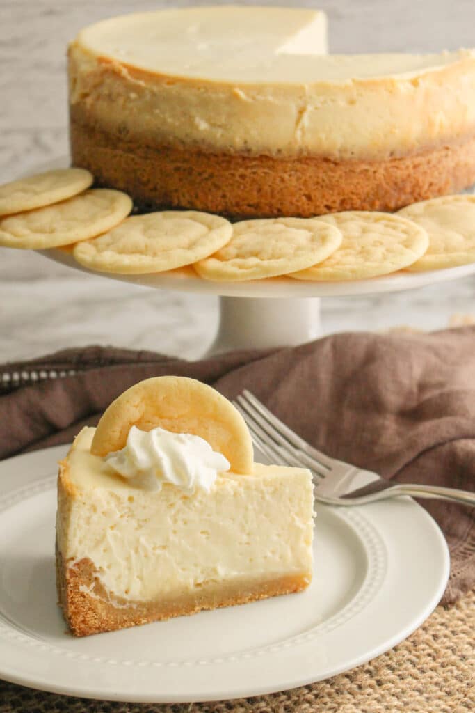 Vertical image of a slice of sugar cookie cheesecake, and a full cheesecake on a white cake stand.