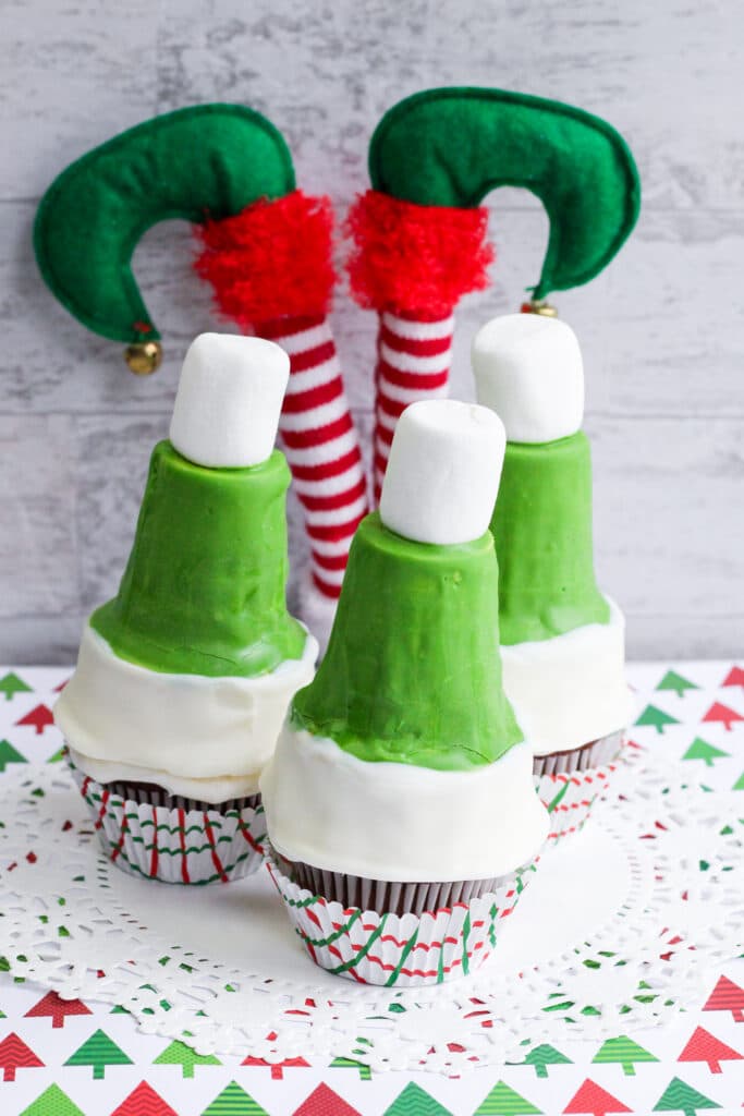 vertical image of three green elf hat cupcakes with a stuffed upside elf behind them