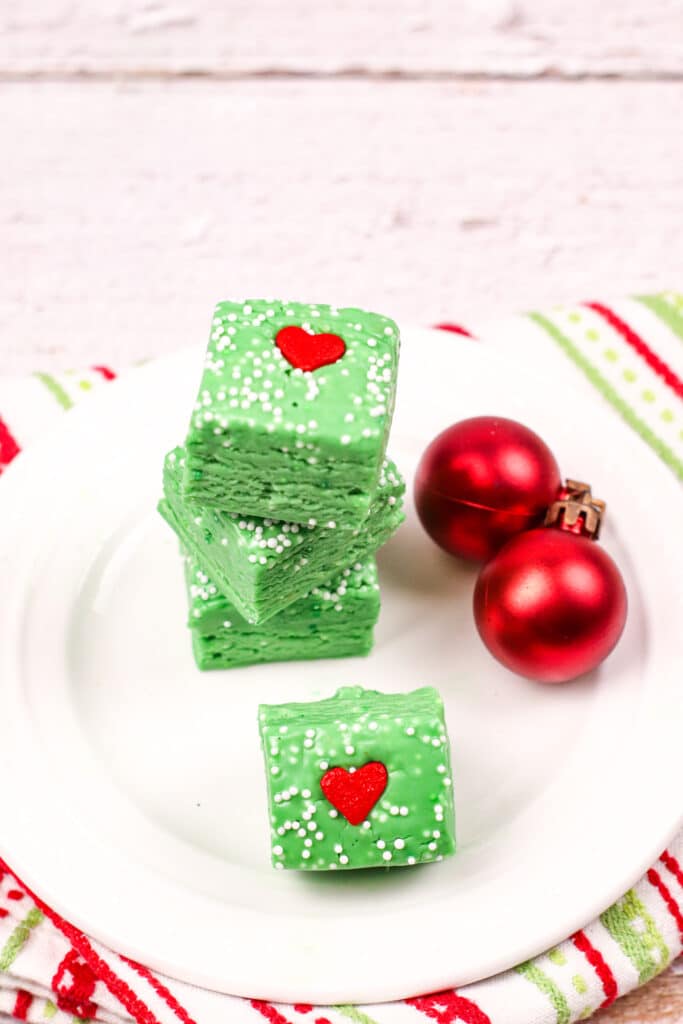 Four pieces of grinch fudge on a white plate with two red christmas ornaments