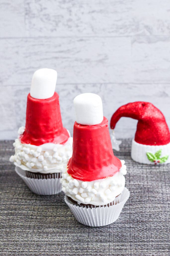 vertical image of two santa hat cupcakes with a fake santa in the background