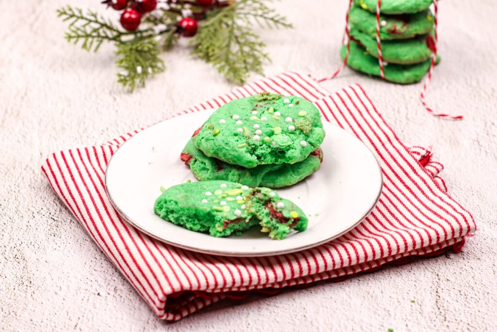 three grinch cake mix cookies on a white plate with one cookie broken in half.