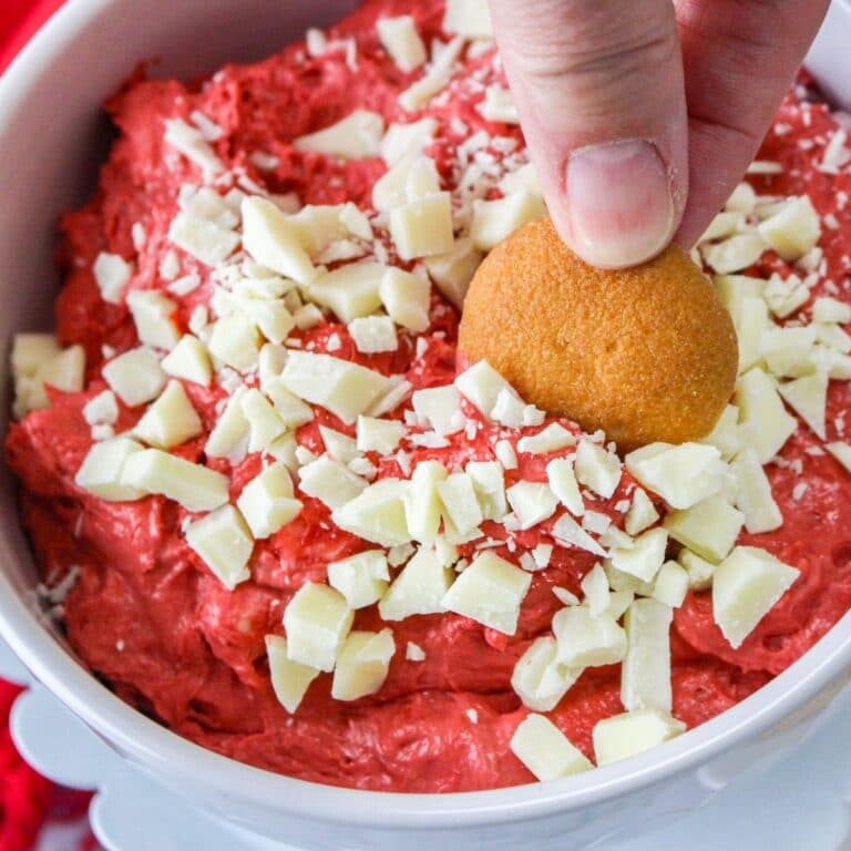 square image of a bowl of red velvet cake batter dip with a womans fingers