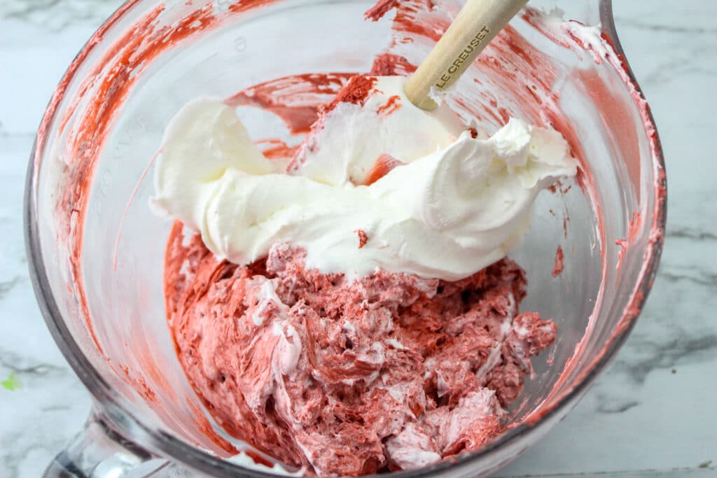 cool whip, red velvet cake mix, cream cheese being mixed in a glass mixing bowl