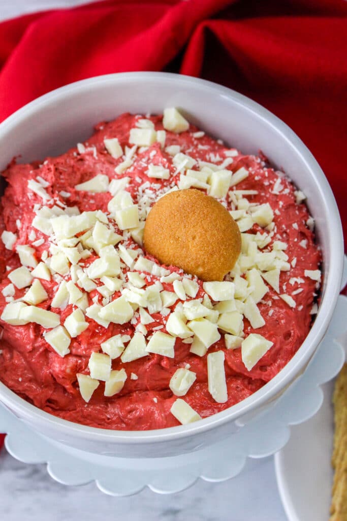 close up birds eye image of white bowl filled with red velvet cake batter dip, topped with chopped white chocolate and a nilla wafer