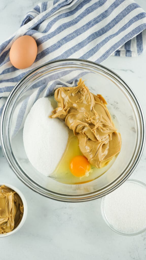peanut butter, sugar and egg in a glass mixing bowl on a marble counter