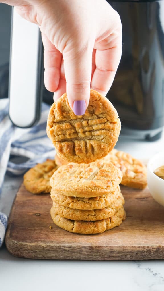 stack of four peanut butter cookies on a wooden cutting board with one cookie held in a womens hand