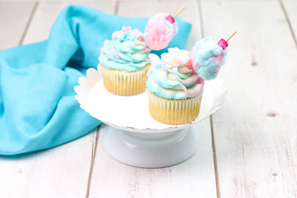 two cotton candy cupcakes on a white cake stand with scalloped edges