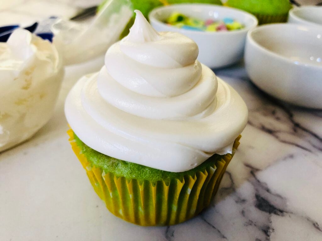 green cupcake being frosted with vanilla frosting on a marble countertop