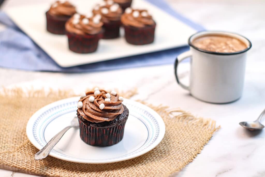 Single hot Cocoa Cupcakes on a white plate with a mug of hot chocolate in the background