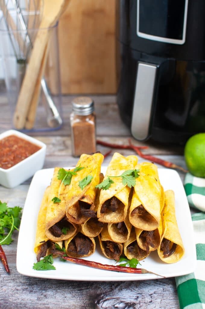 vertical image of air fryer beef Taquitos on a white plate with an air fryer in the background