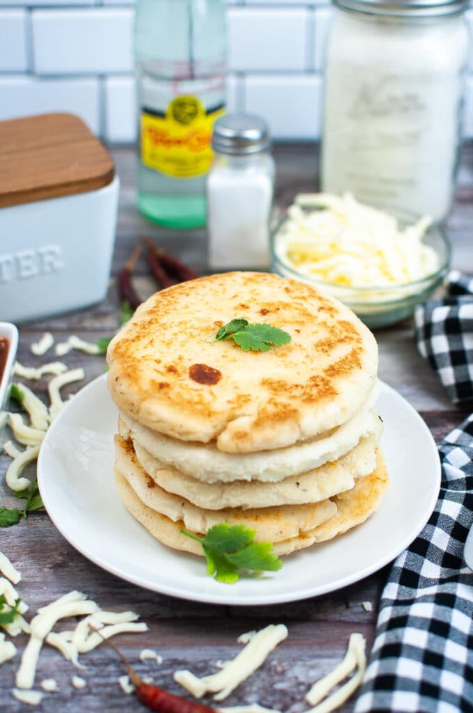 vertical image of arepas con queso on a white plate with a small bowl of shredded cheese in the background