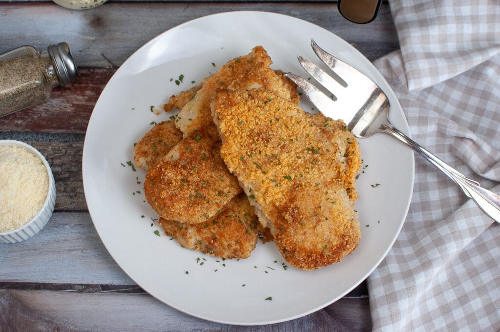 birds eye image of three air fryer parmesan crusted chicken breasts on a white plate with a large serving fork