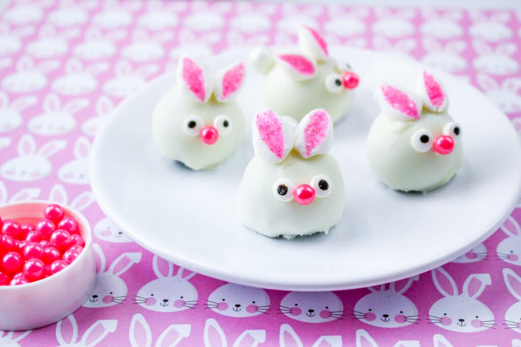 four bunny oreo balls on a white plate with a purple bunny tablecloth underneath
