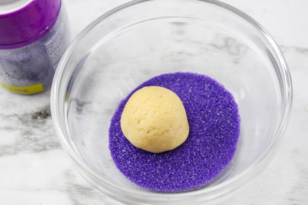 ball of cookie dough being rolled in purple sanding sugar
