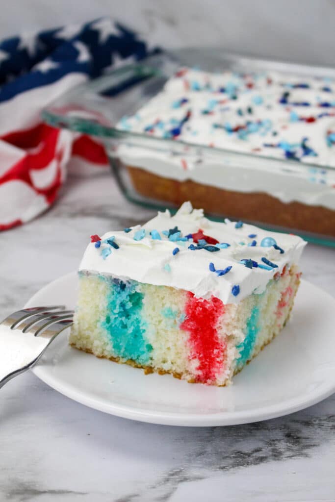 a slice of 4th of july poke cake on a white plate with the whole cake in the background