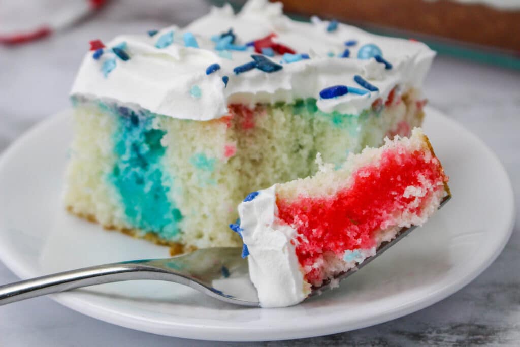 Close up of a slice of 4th of July Poke Cake on a white plate with a bite on a fork