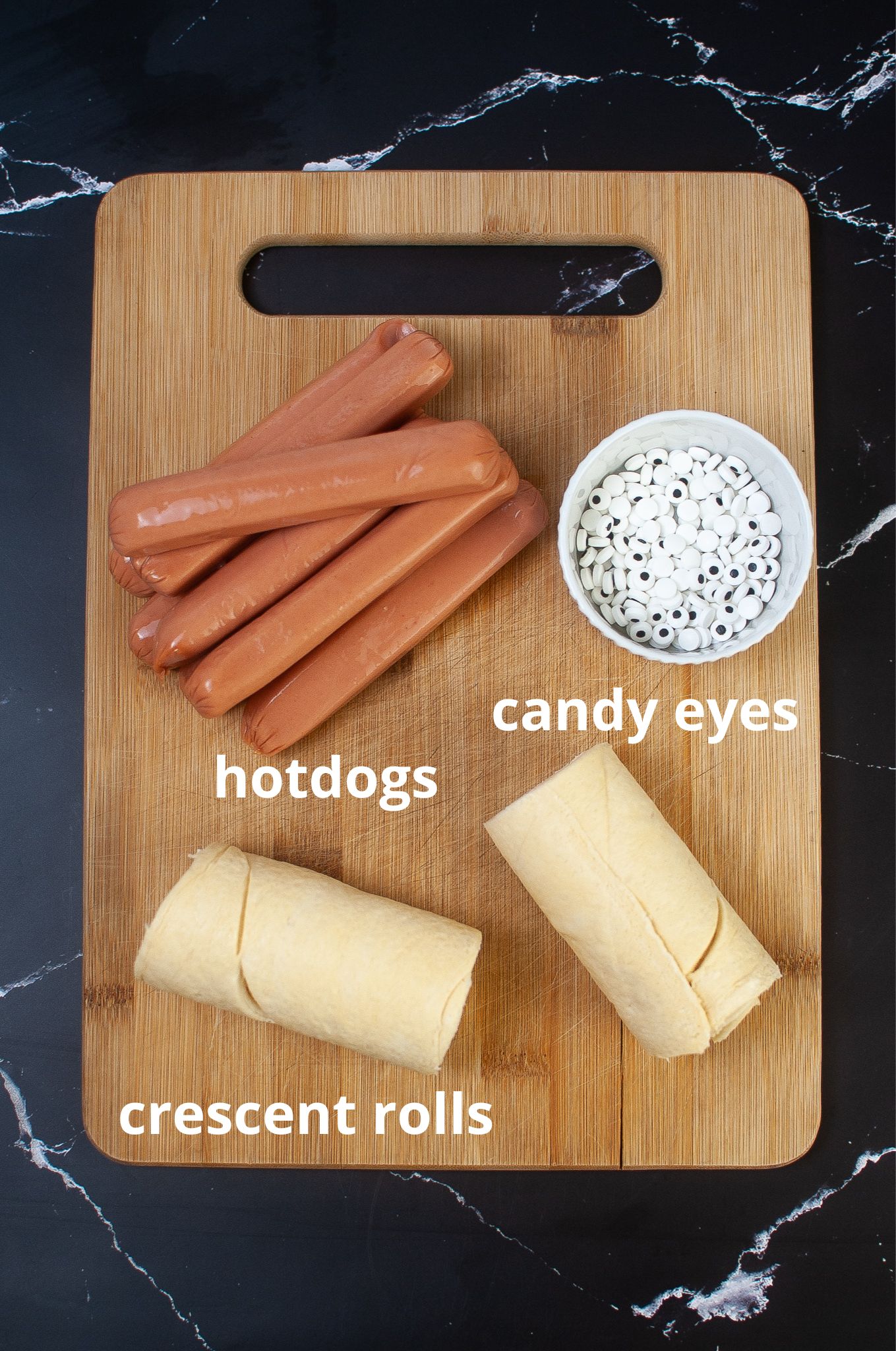 hotdogs, candy eyeballs, and cresecent rolls on a wooden cutting board