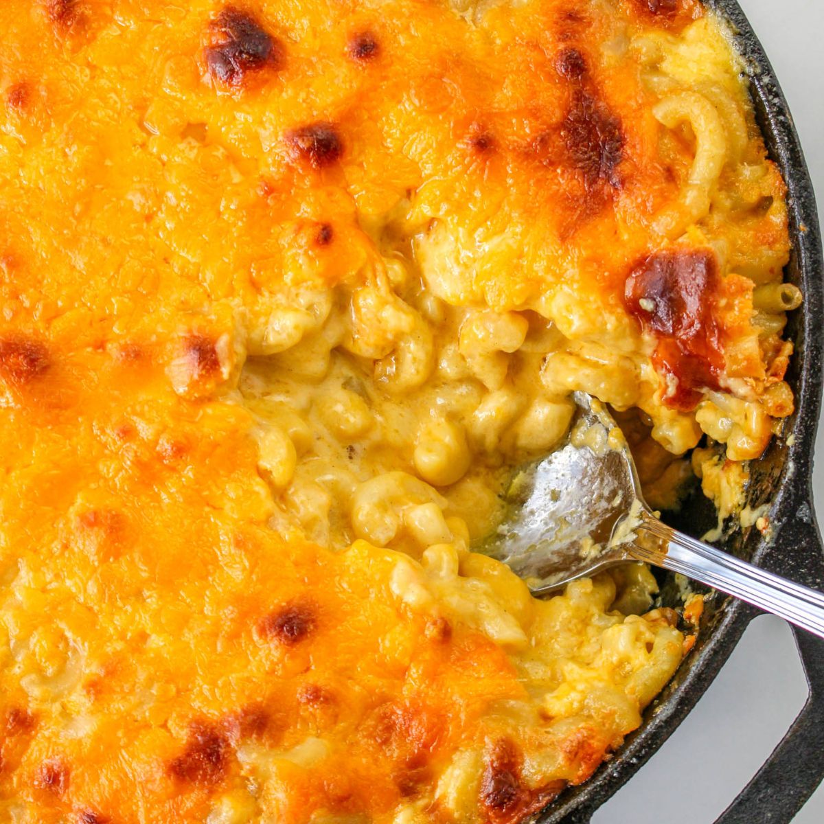 Baked cast iron skillet mac and cheese with a golden, melted cheese topping, served hot as a metal spoon scoops out a delicious portion.