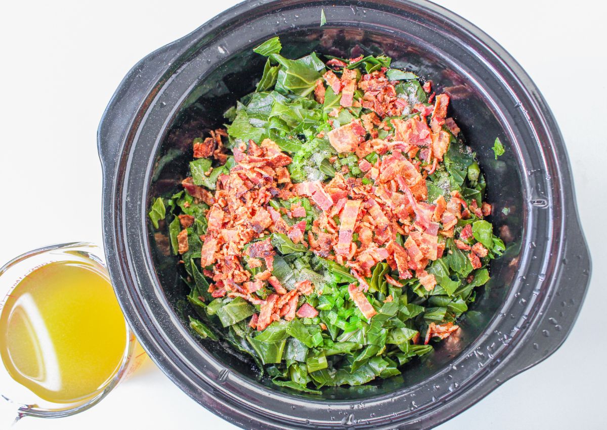 bacon and collard greens in a slow cooker with a measuring cup of chicken broth next to it