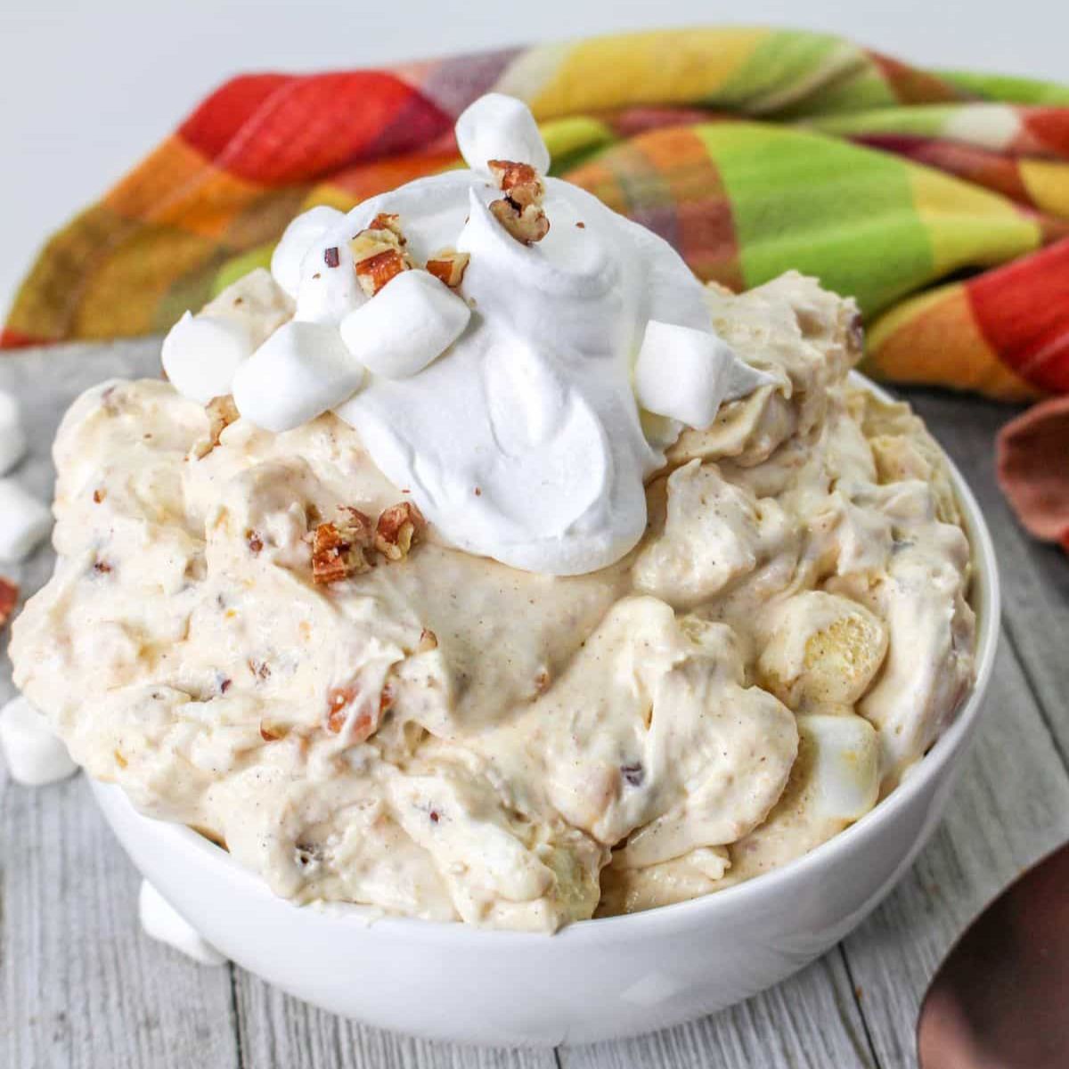 A bowl of creamy Sweet Potato Fluff dessert topped with whipped cream, mini marshmallows, and chopped nuts sits on a wooden surface.
