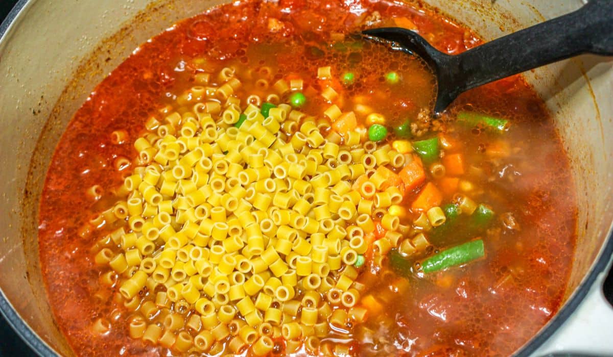 pasta and frozen vegetables being added to a dutch oven filled with soup