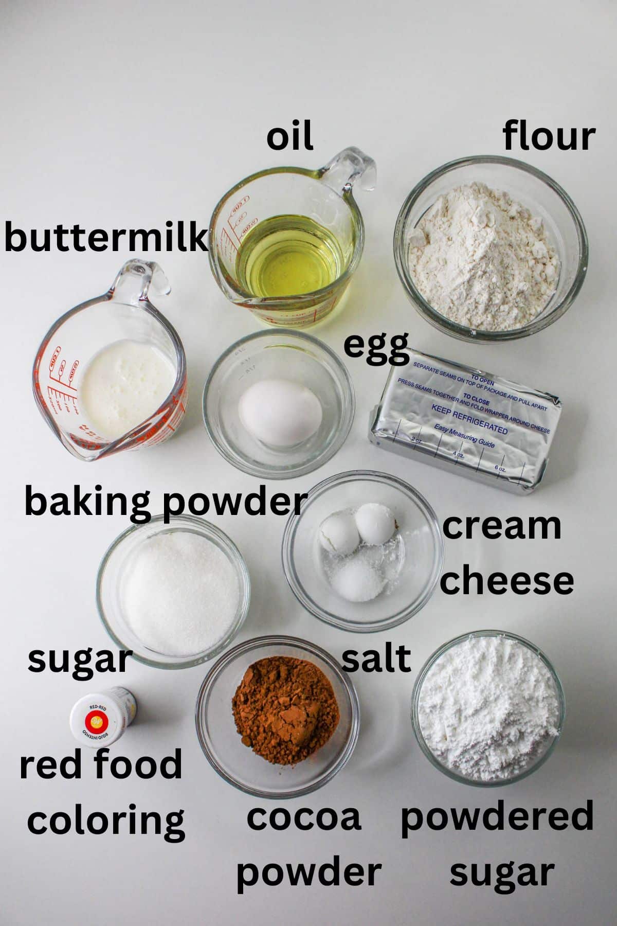 buttermilk, oil, flour, egg, baking powder, cream cheese, sugar, salt, red food coloring, cocoa powder, and powdered sugar on a white background